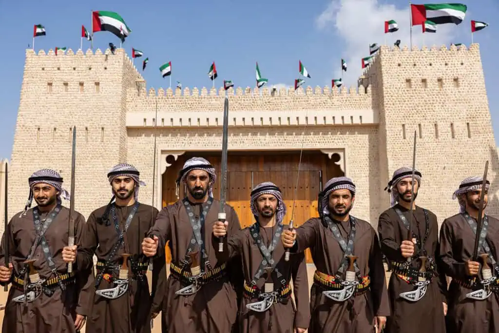 Traditional Emirati performers in front of a historic fortress with flags.